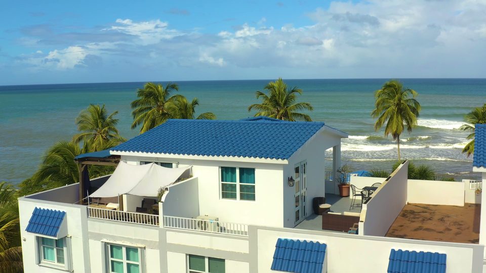 The penthouse rooftop from above: the blue-tile roof of the rooftop room, white walls, the shaded lounge and terrace, palms and the open Atlantic behind