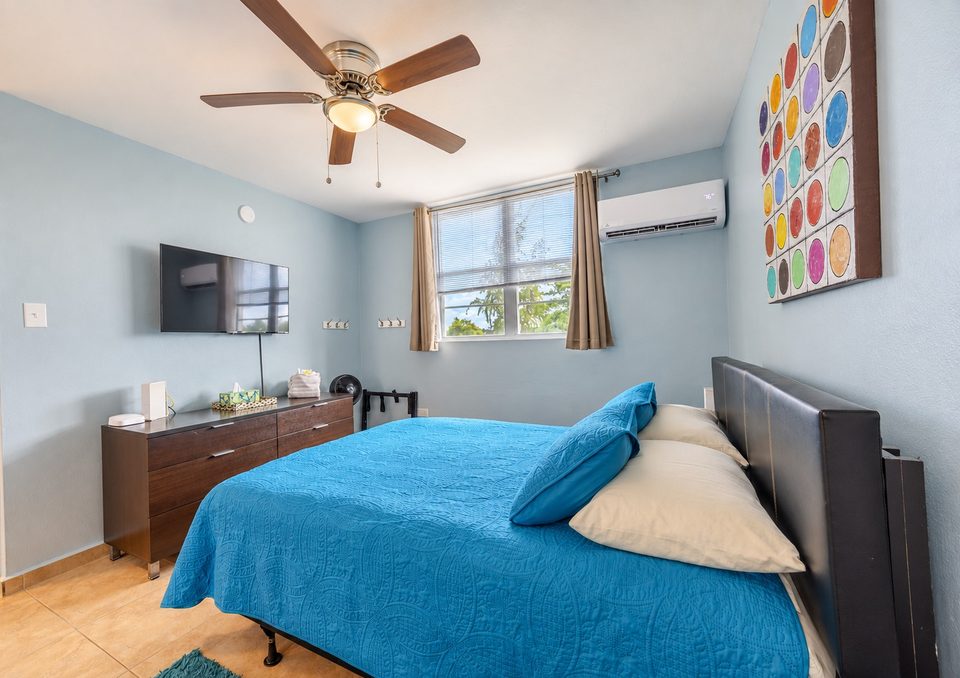 Bedroom two: a bed with a bright blue spread, wall-mounted TV over the dresser, ceiling fan, ductless air conditioning and a window