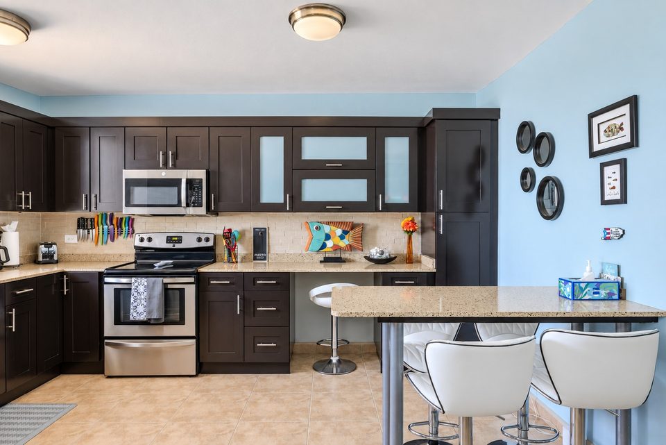 The whole kitchen at once: range, microwave, dark cabinets and the breakfast bar with two white stools