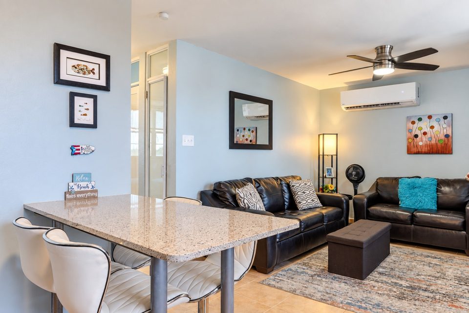 From the breakfast bar into the living room: black leather sofas, a ceiling fan, ductless air conditioning and framed art