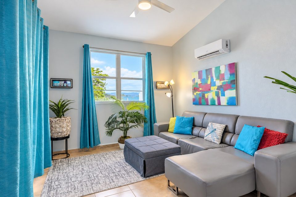 The grey sectional under turquoise curtains, a window with a slice of ocean, a floor lamp and a plant
