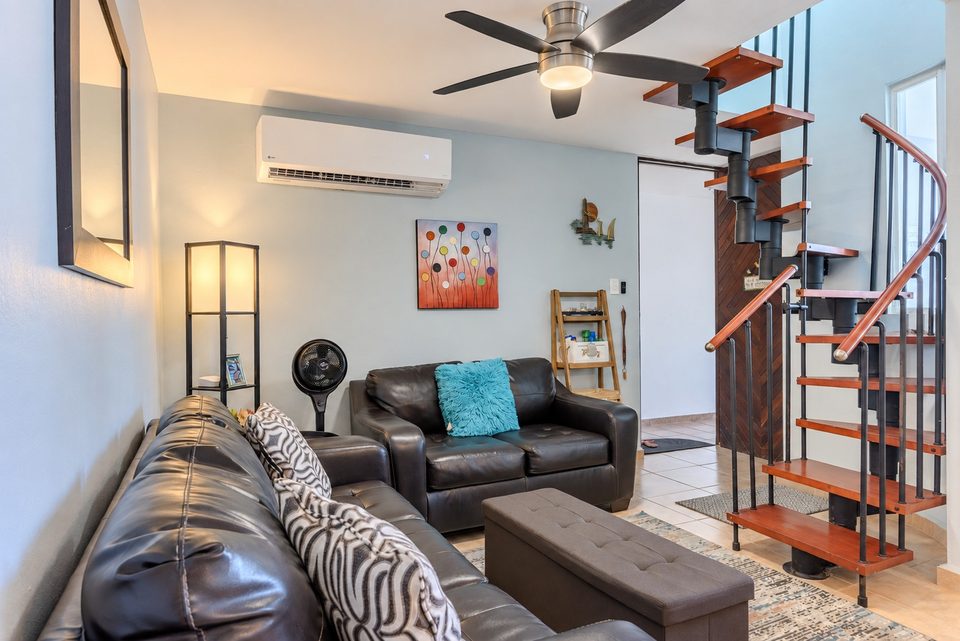 Black leather sofas and an ottoman beside the red-and-black spiral stair to the loft; ceiling fan and ductless air conditioning above