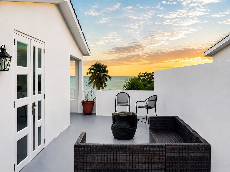 The private rooftop terrace: white French doors, wicker lounge seating, two chairs by the wall, palms and the open Atlantic beyond